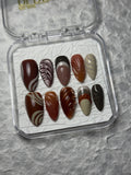 A set of 10 false press-on nails in a clear plastic case, featuring various designs including solid colors, stripes, and swirls in shades of brown, beige, and black, displayed on a textured gray background.