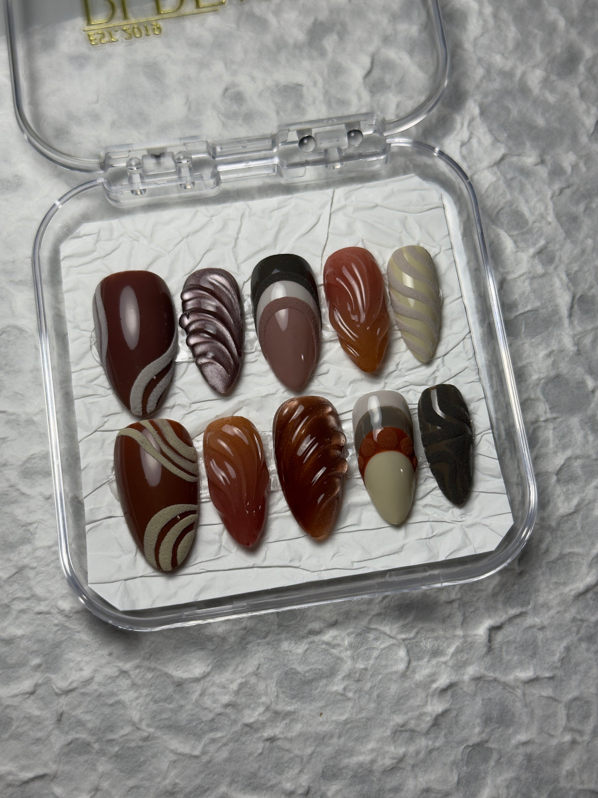 A set of 10 false press-on nails in a clear plastic case, featuring various designs including solid colors, stripes, and swirls in shades of brown, beige, and black, displayed on a textured gray background.