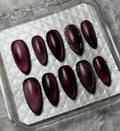 A set of ten glossy dark burgundy press-on nails with thin gold accent lines, displayed in a clear plastic case on a textured gray background. The brand name 'PI BEAUTY NAILS