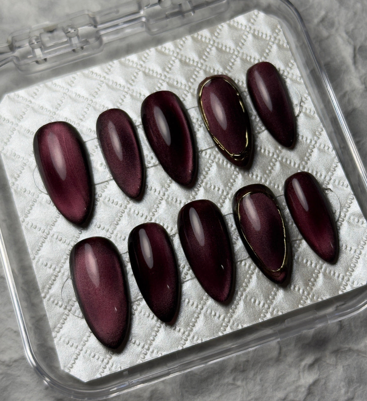 A set of ten glossy dark burgundy press-on nails with thin gold accent lines, displayed in a clear plastic case on a textured gray background. The brand name 'PI BEAUTY NAILS