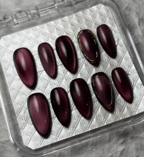 A set of ten glossy dark burgundy press-on nails with thin gold accent lines, displayed in a clear plastic case on a textured gray background. The brand name 'PI BEAUTY NAILS