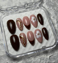 A set of 10 press-on false nails in a clear plastic case, featuring designs of solid dark brown, light pink with gold leaf accents, and light pink with white floral details, on a textured gray background.