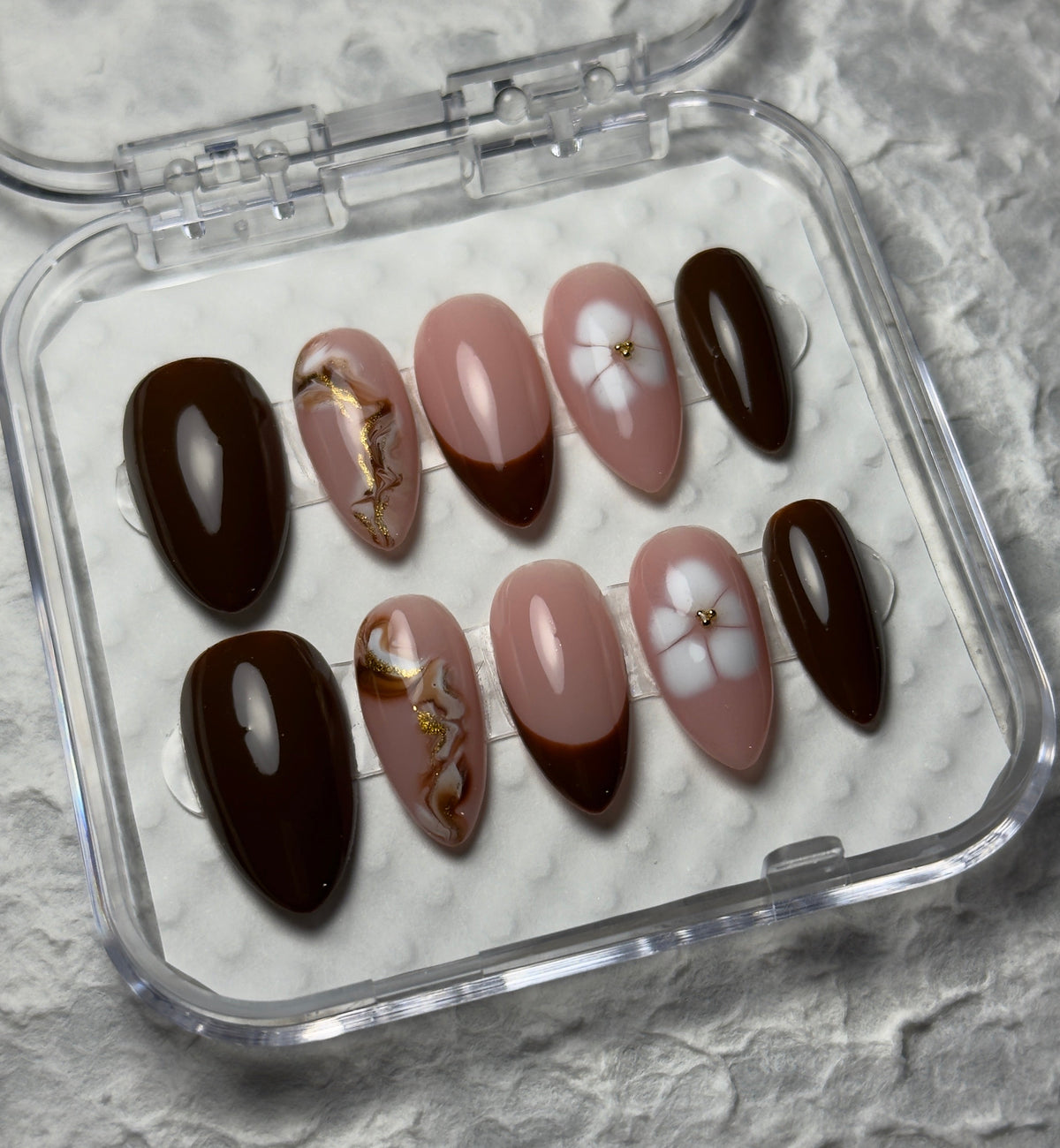 A set of 10 press-on false nails in a clear plastic case, featuring designs of solid dark brown, light pink with gold leaf accents, and light pink with white floral details, on a textured gray background.