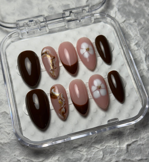 A set of 10 press-on false nails in a clear plastic case, featuring designs of solid dark brown, light pink with gold leaf accents, and light pink with white floral details, on a textured gray background.