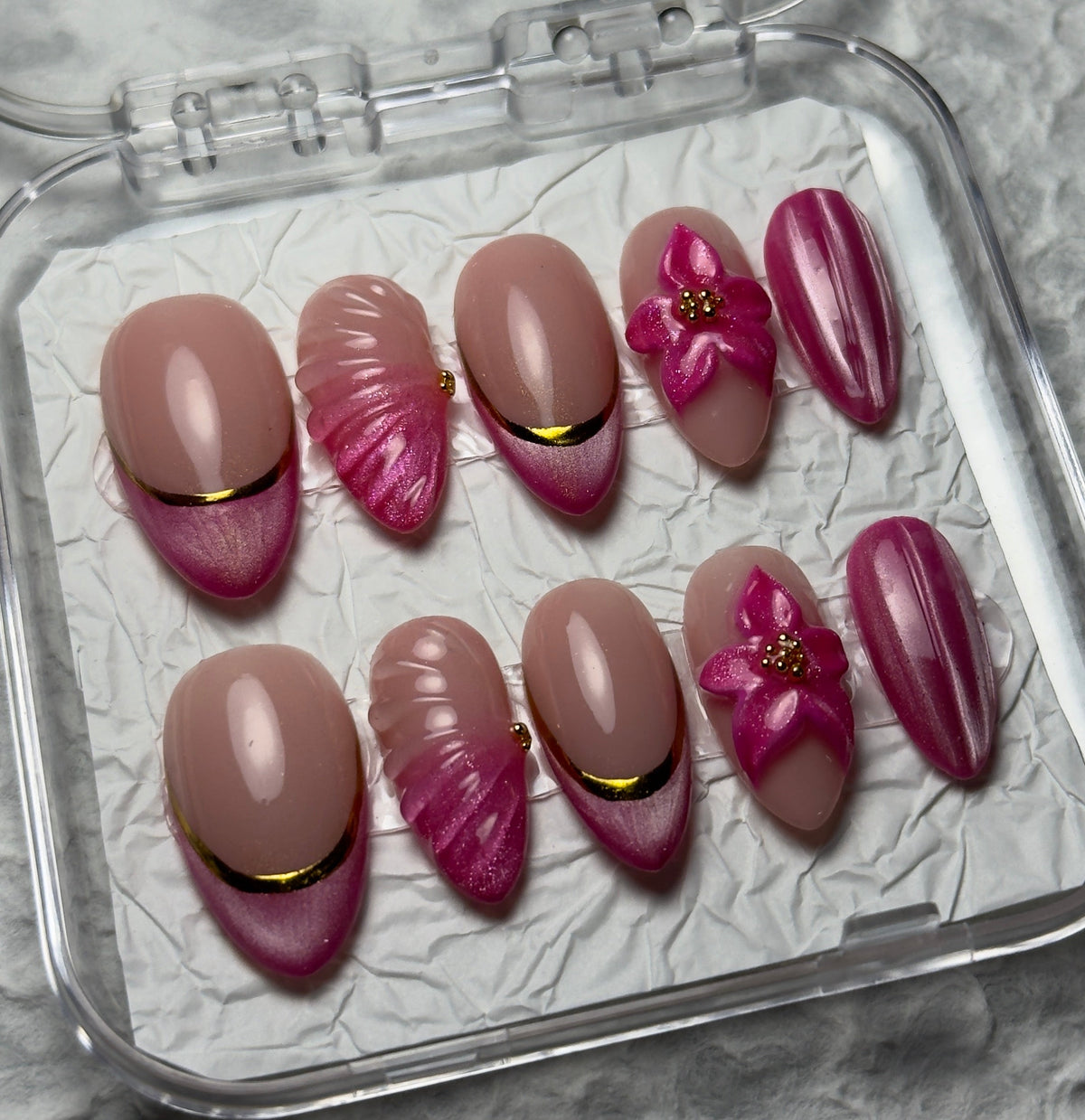 A set of 10 false press-on nails in a clear plastic case, featuring a mix of nude pink, magenta, and metallic gold designs, with the brand name 'BI BEAUTY' visible on the inside lid.