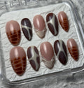 A set of 10 press-on nails displayed in a clear plastic case on a textured gray background. The nails feature a mix of nude pink, dark brown, and orange-brown patterns, including stripes, swirls, and feather-like designs.