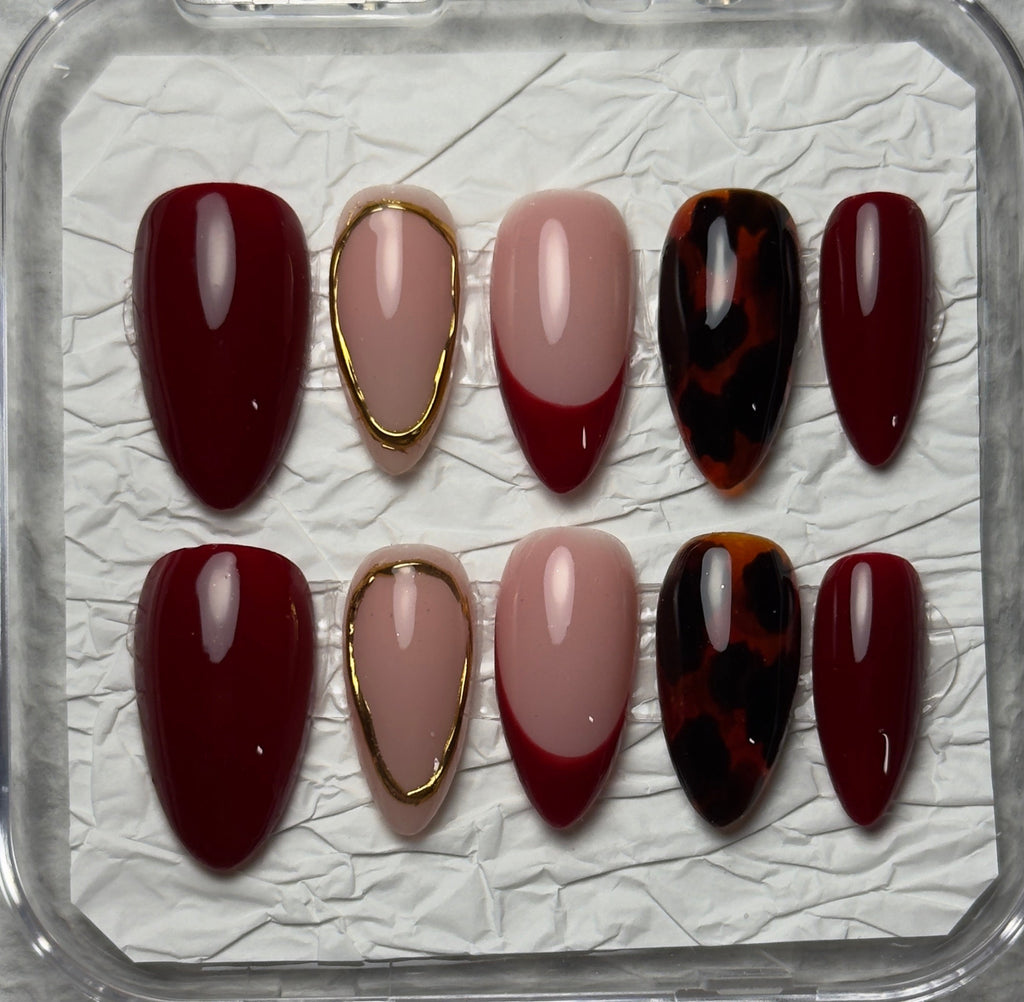 A set of 10 press-on nails displayed in a clear plastic case on textured white paper. The nails feature a mix of deep burgundy, dusty pink, metallic gold trim, and a dark red and black tortoiseshell pattern.