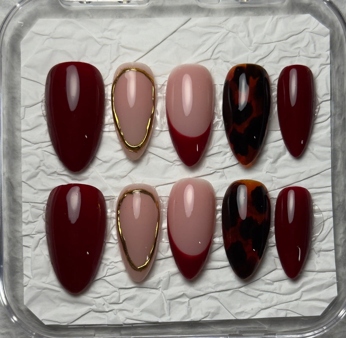 A set of 10 press-on nails displayed in a clear plastic case on textured white paper. The nails feature a mix of deep burgundy, dusty pink, metallic gold trim, and a dark red and black tortoiseshell pattern.