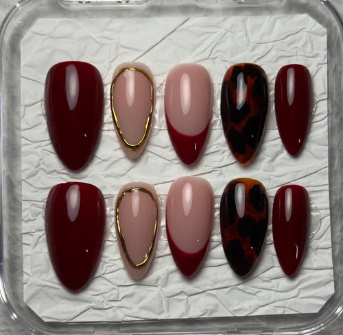 A set of 10 press-on nails displayed in a clear plastic case on textured white paper. The nails feature a mix of deep burgundy, dusty pink, metallic gold trim, and a dark red and black tortoiseshell pattern.