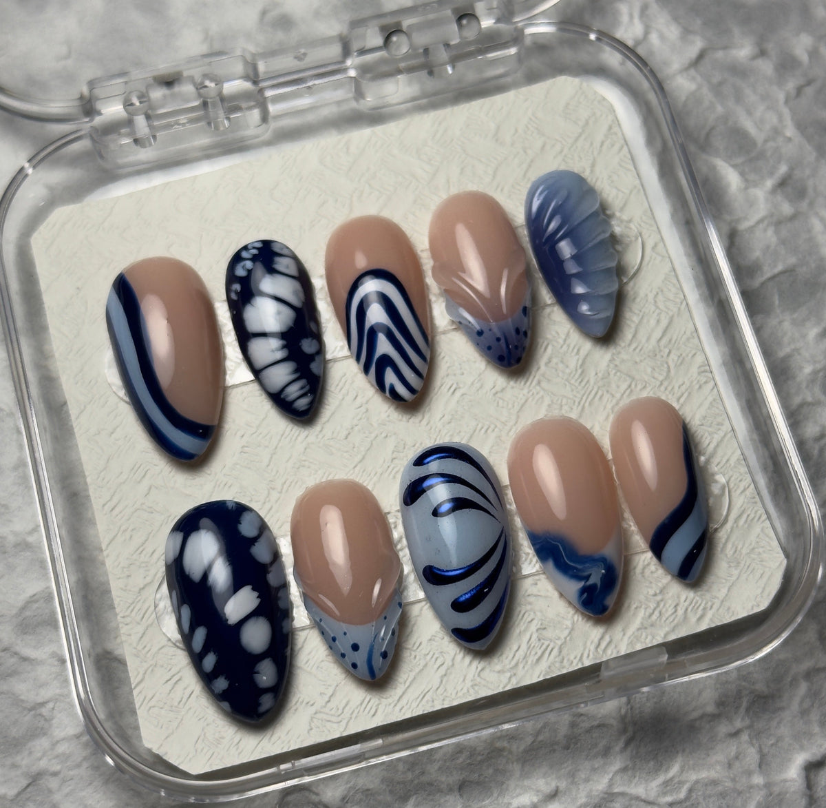 A set of 10 press-on nails displayed in a clear plastic case on a textured gray surface. The nails feature a mix of nude beige, navy blue, and white patterns including stripes, dots, and abstract swirls.